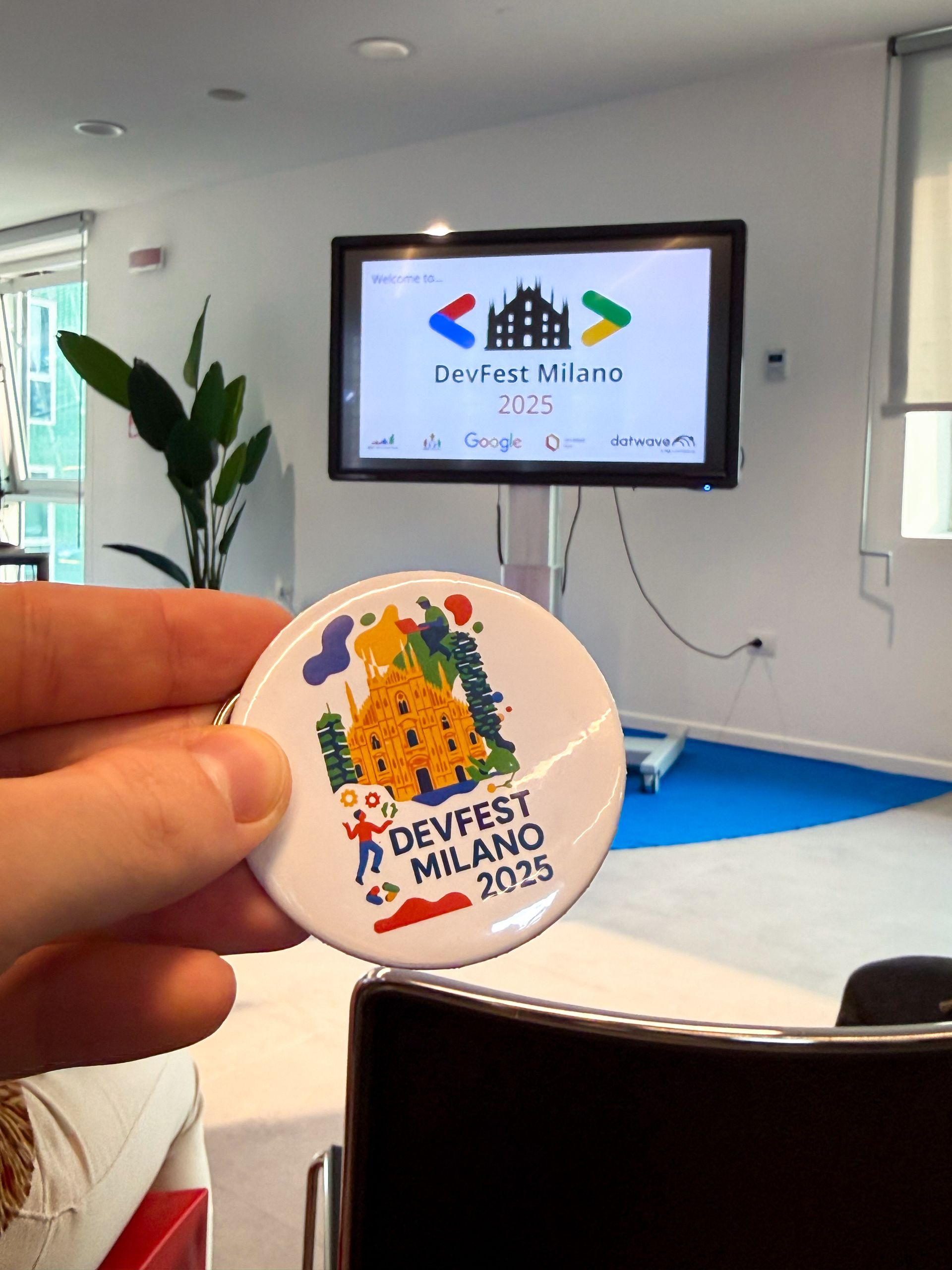 The round DevFest Milano 2025 pin badge held up in front of the screen