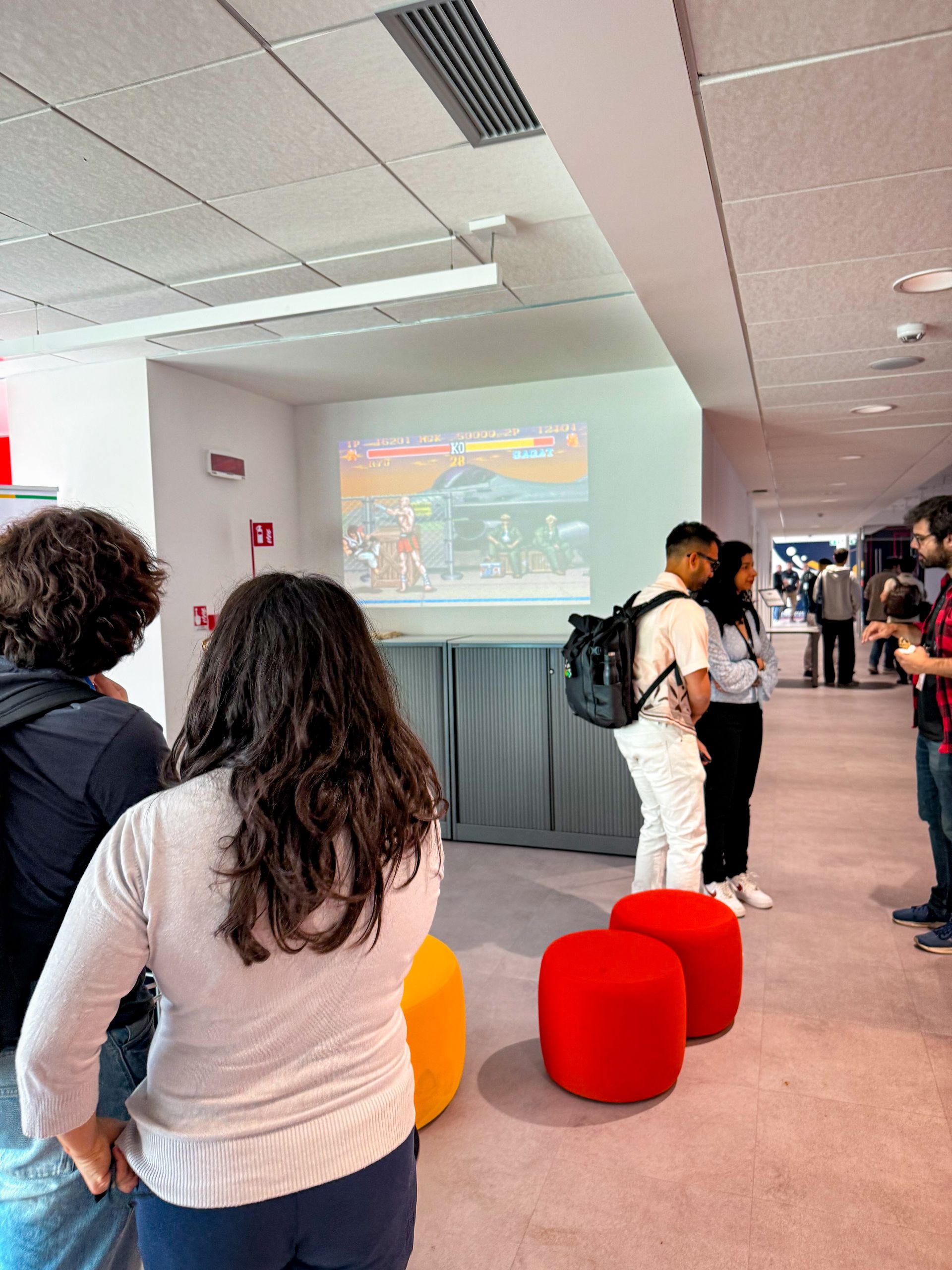 The foyer of the venue with orange and red poufs and people watching a projection
