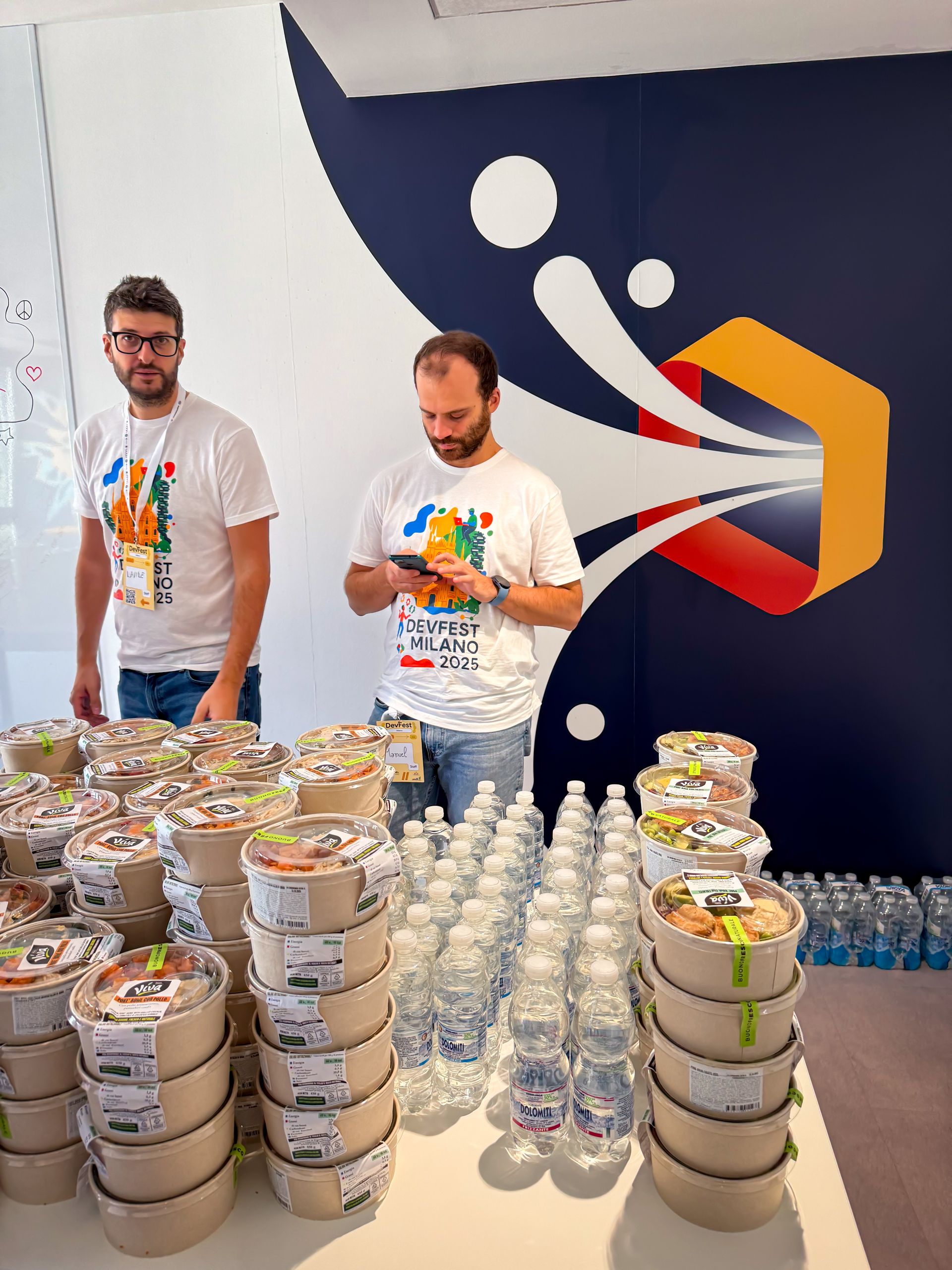 Volunteers in DevFest t-shirts behind a table of lunch boxes and water bottles