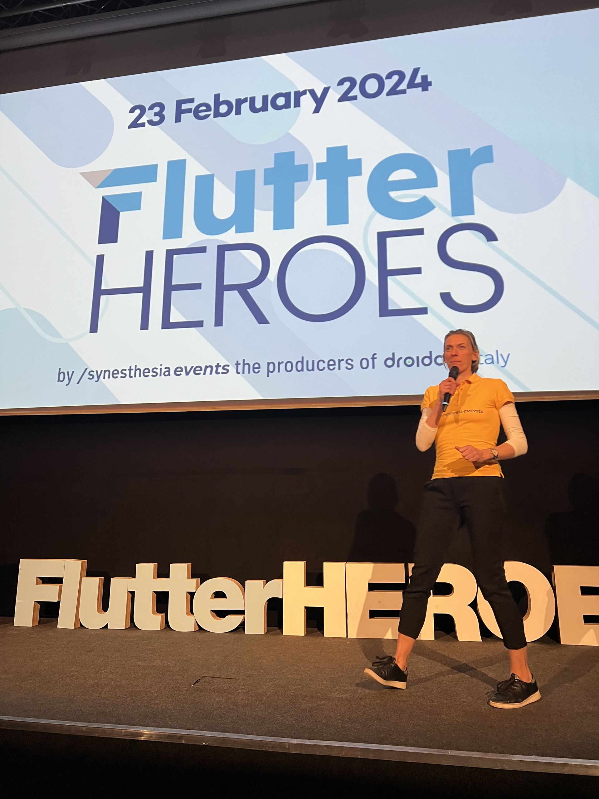 A host in an orange synesthesia events shirt opening Flutter Heroes 2024 on stage