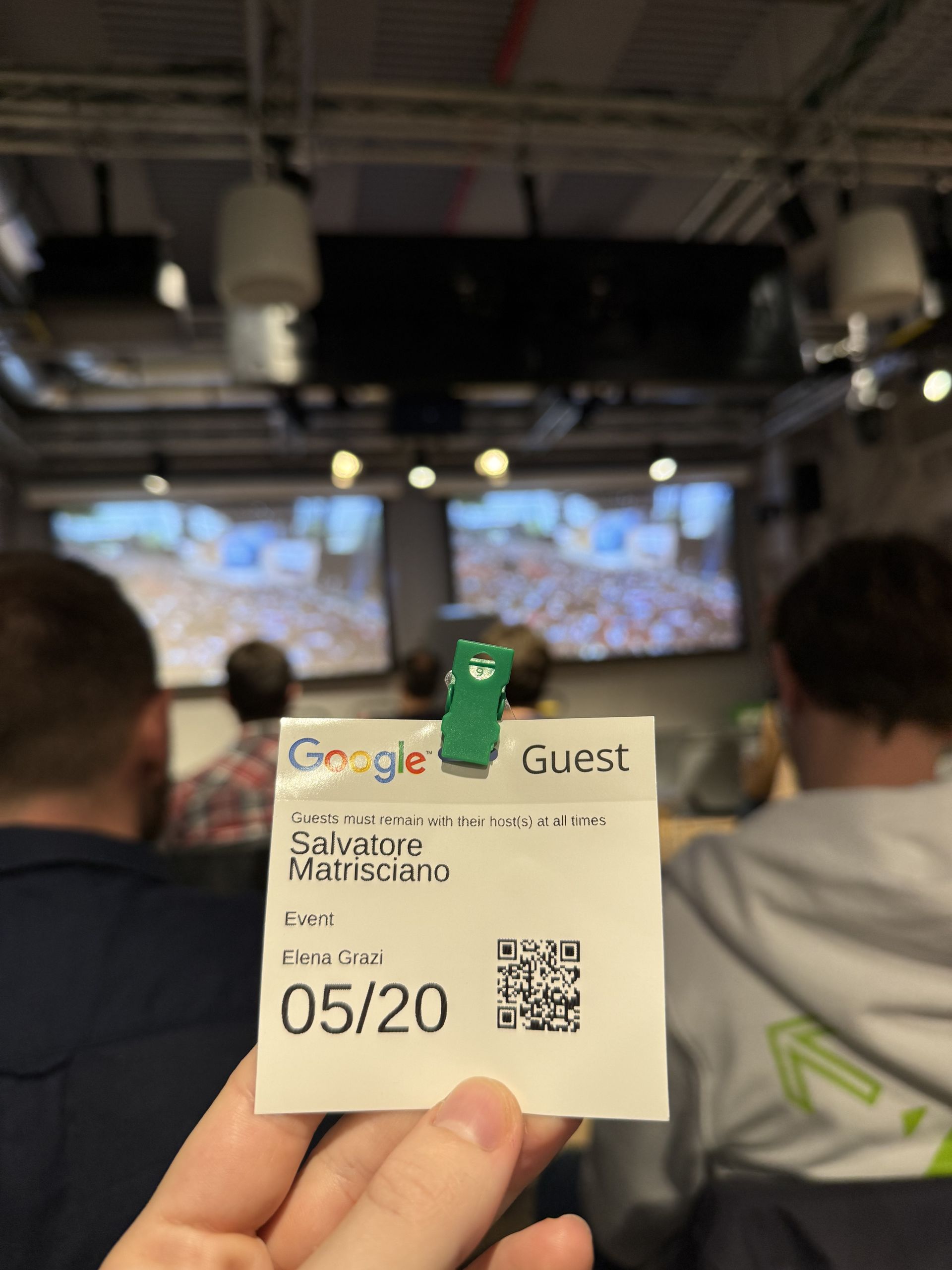 A Google Guest badge with the name and the date 05/20 held up in front of the screens