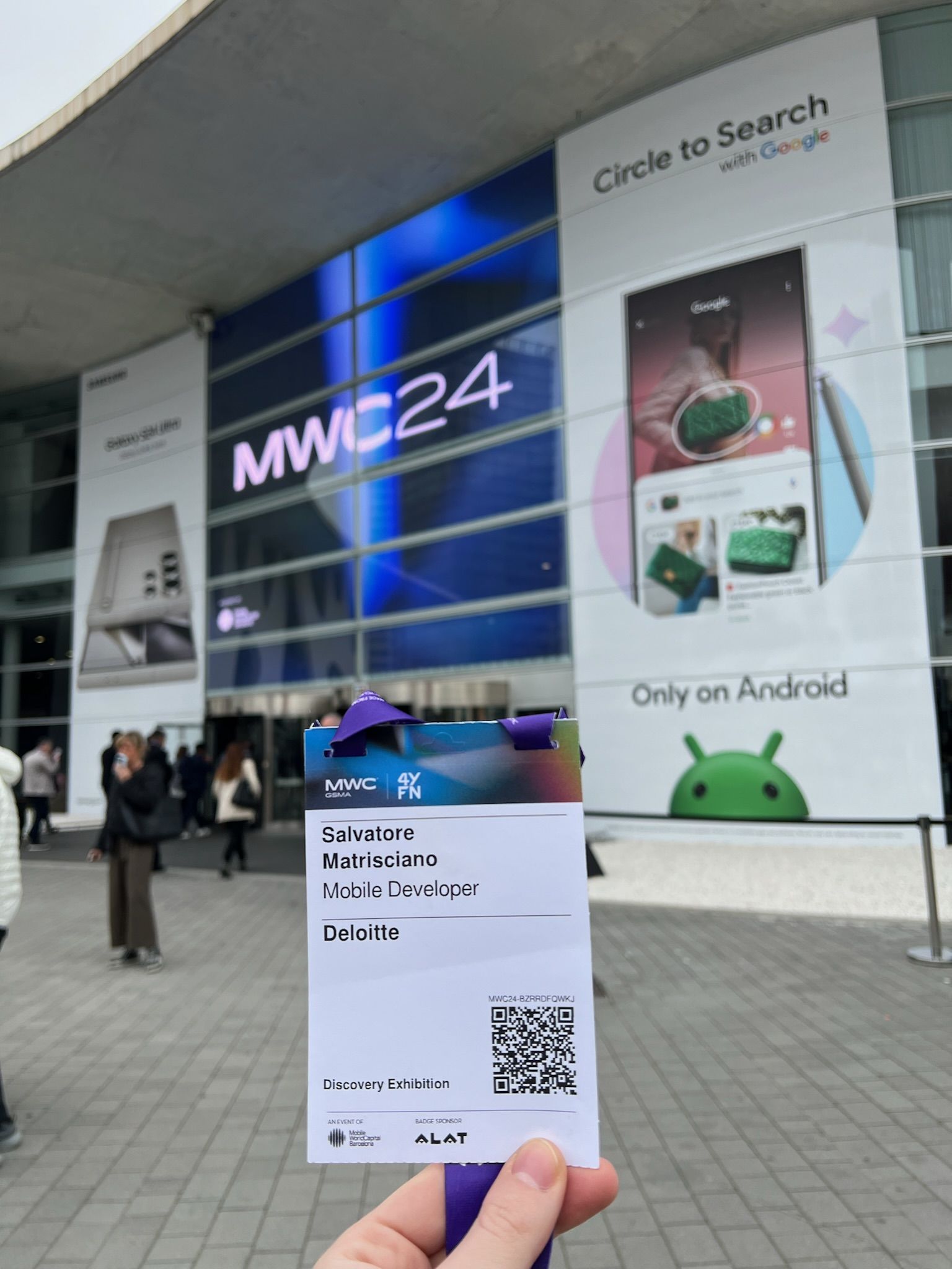 The MWC24 badge held up in front of the Fira Gran Via facade