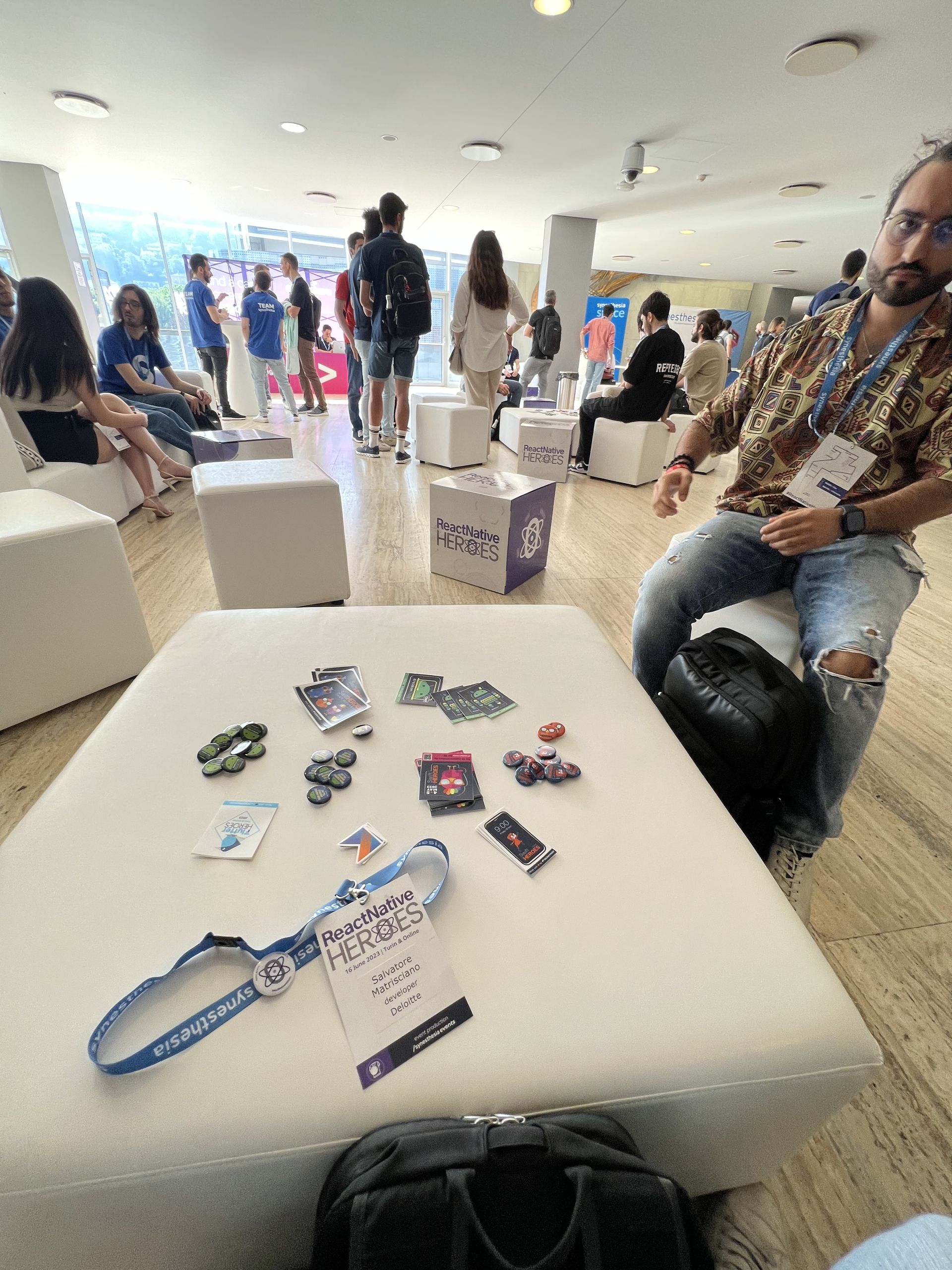 The React Native Heroes badge and lanyard on a pouf, surrounded by stickers and pins