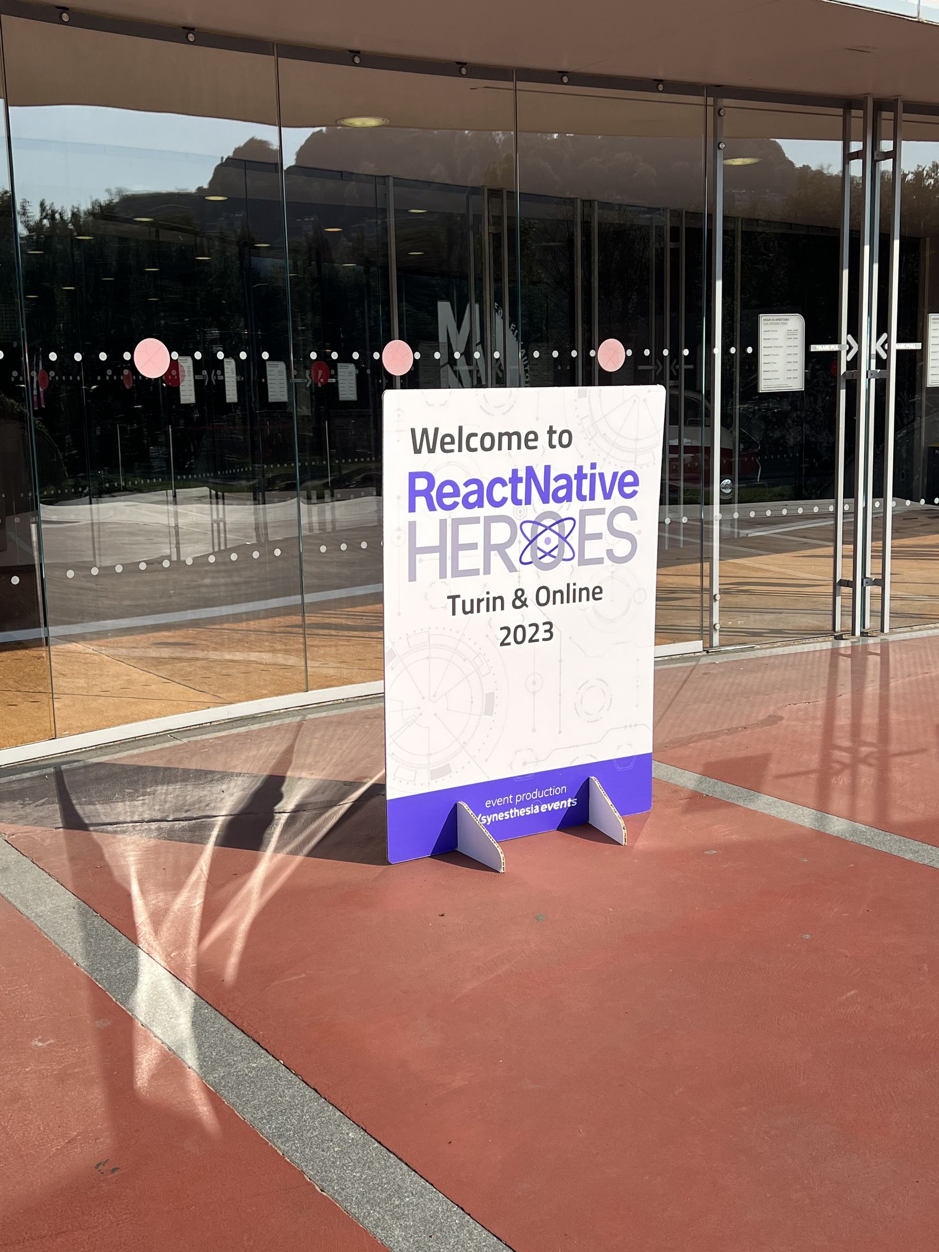The Welcome to ReactNative Heroes Turin and Online 2023 sign outside the glass doors of the MAUTO